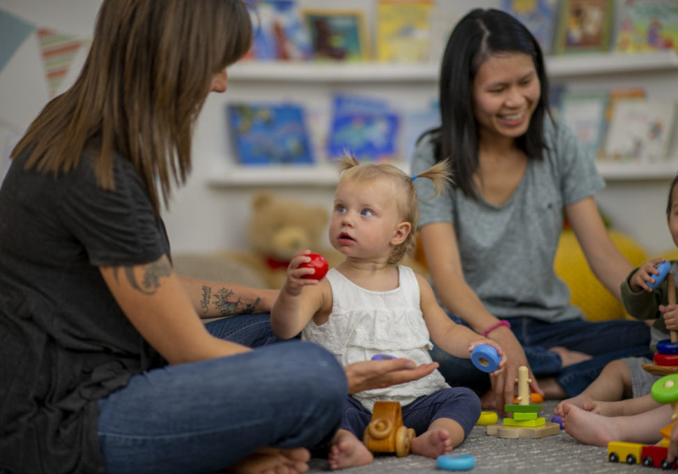 A multi-ethnic group of moms sit in a circle as their babies play in a nursery
